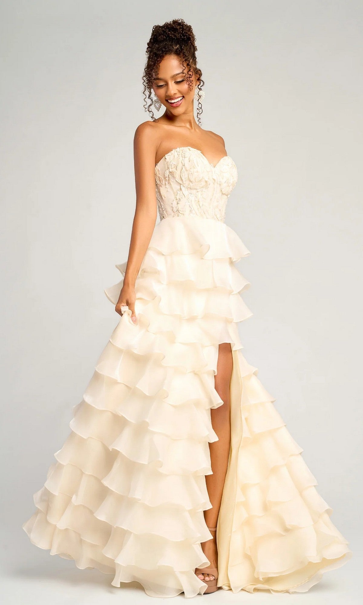 A woman models the Portia and Scarlett PS26048 long formal dress in cream, featuring a flowy embroidered corset bodice, ruffled tiered skirt, and high slit, standing before a plain background.
